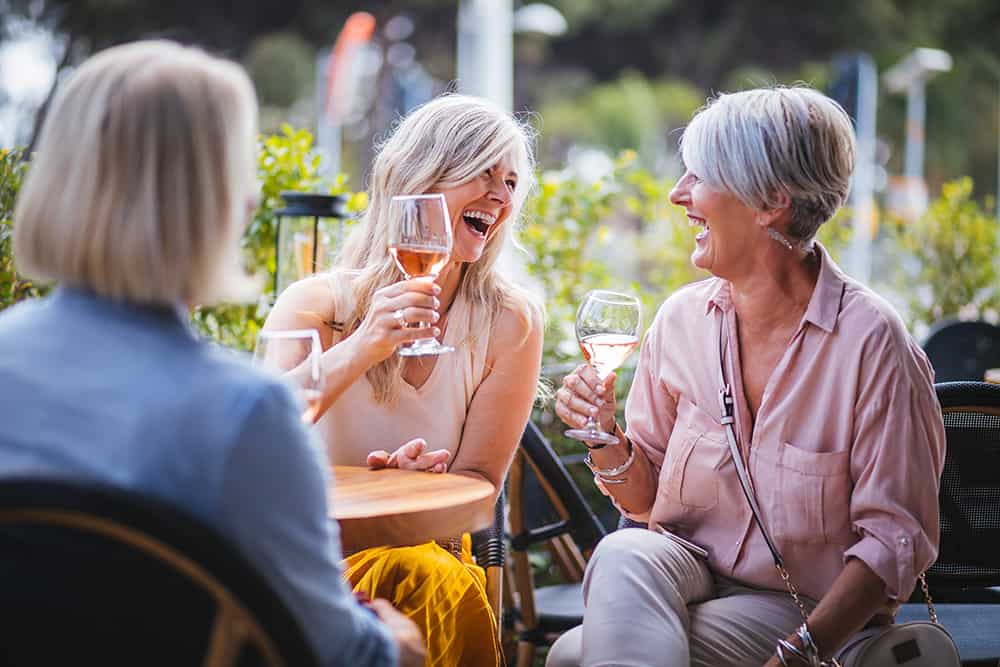 Residents sitting outside with glasses of wine and laughing at Cadence Creek at Gosling 55 and over apartments.